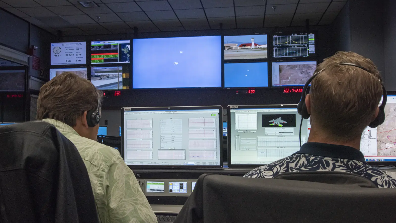 Two engineers with headsets watching flight data on the screens of a flight control room